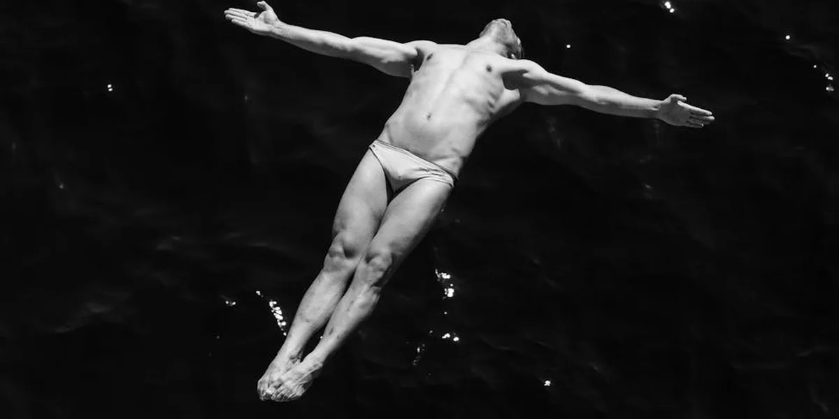 A shirtless man in swim briefs is suspended mid-air with arms outstretched against a dark background, suggesting a fall or descent.