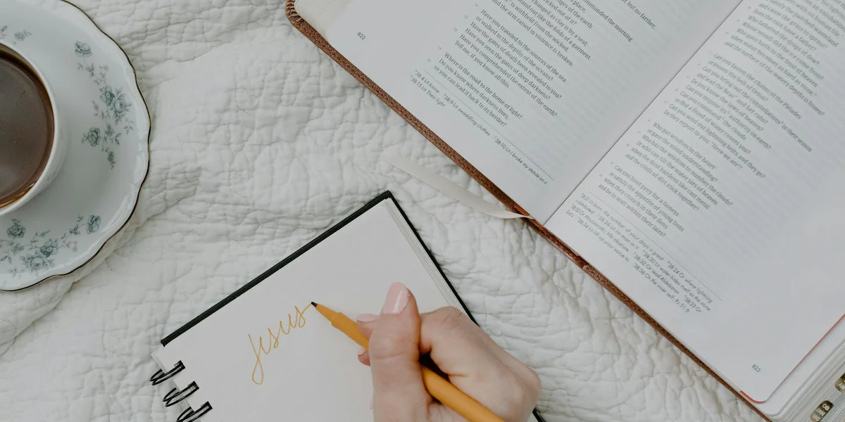 Close-up of a hand writing the word 'dream' on a notebook, with an open book and a cup of coffee on a white quilted surface.