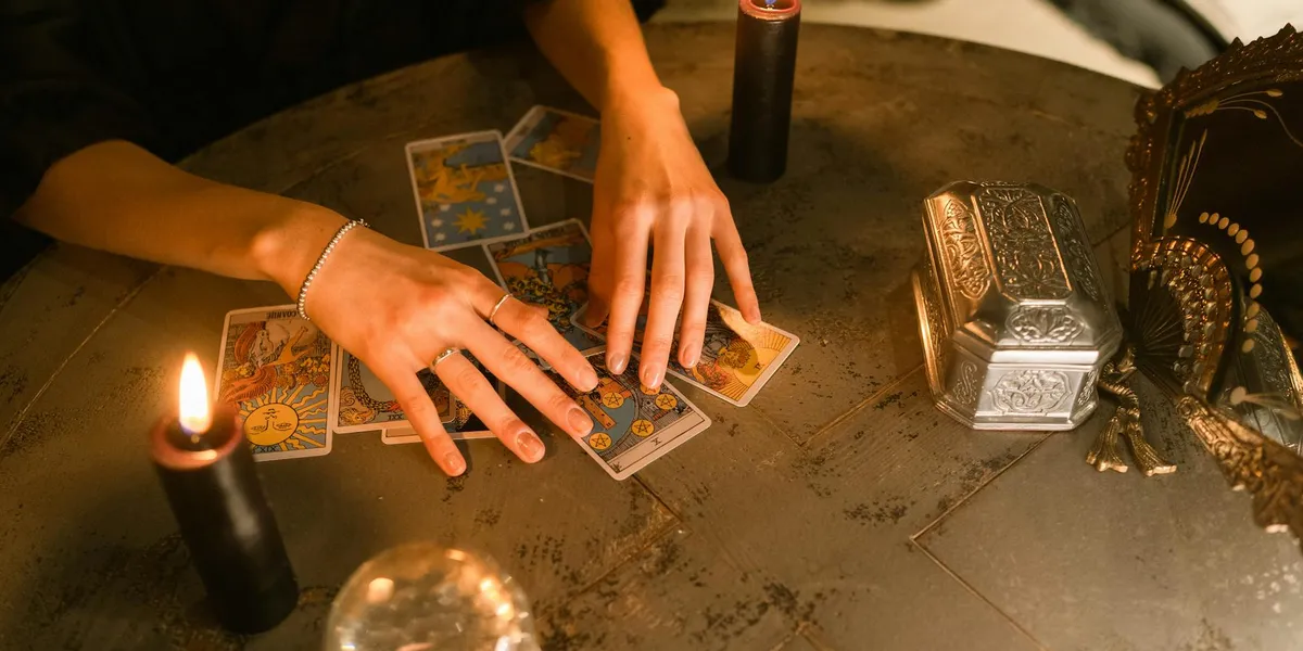 Hands lay out tarot cards on a round, worn table with two lit candles, a metal box, and a beaded bracelet, evoking divination and dream interpretation.