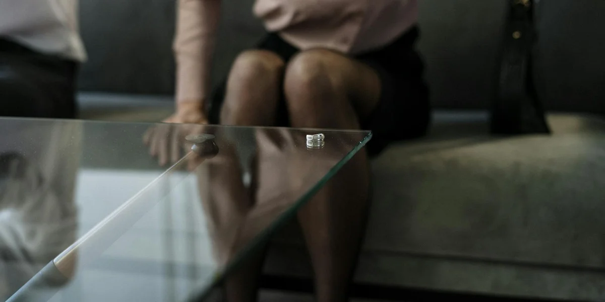 Two people sit on a couch with a glass coffee table in front; a small ring sits on the table, suggesting relationship tension and contemplation.