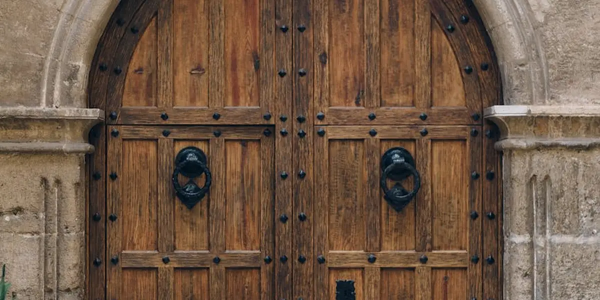 A pair of heavy wooden double doors with iron studs and round door knockers, set in a stone archway.