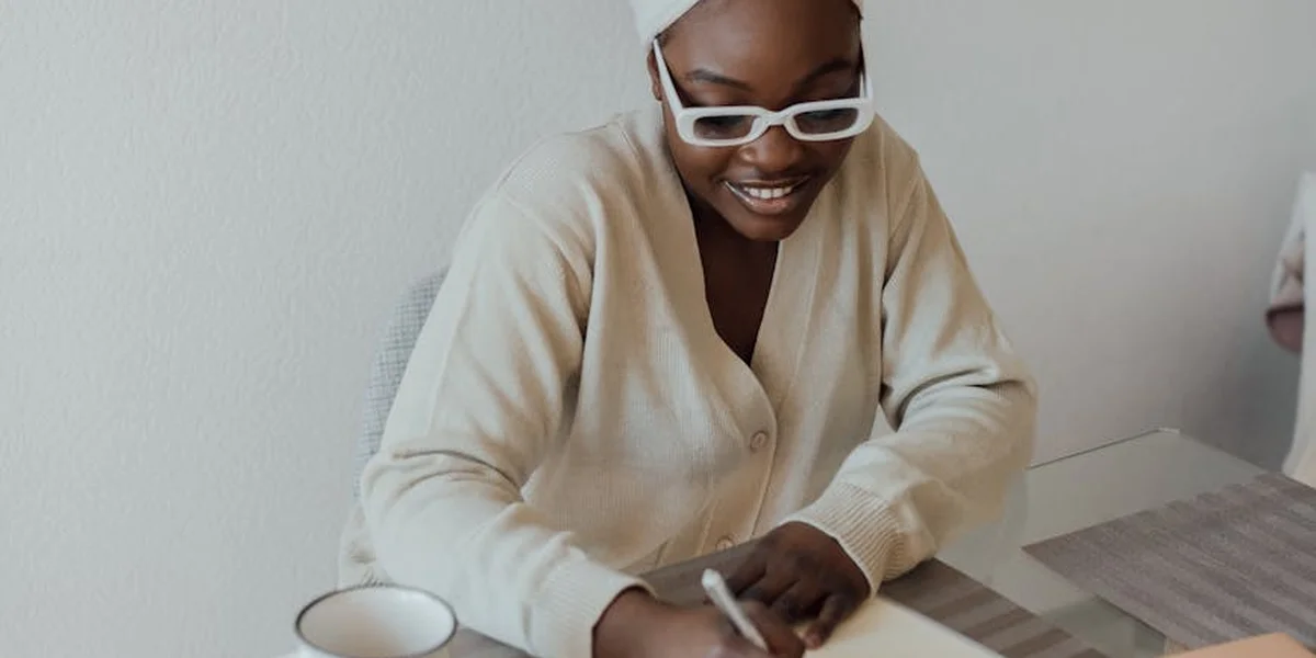 Person wearing glasses sits at a desk, smiling while writing in a notebook, conveying focus on ongoing dream work.