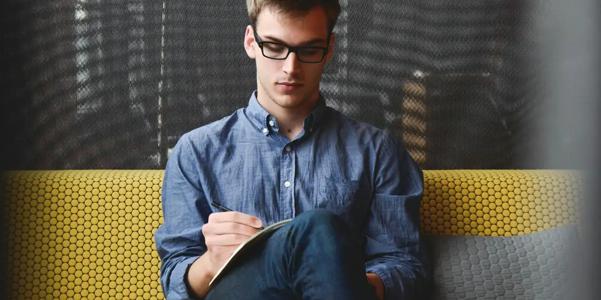 Young man with glasses sits on a yellow patterned couch, reading a notebook.