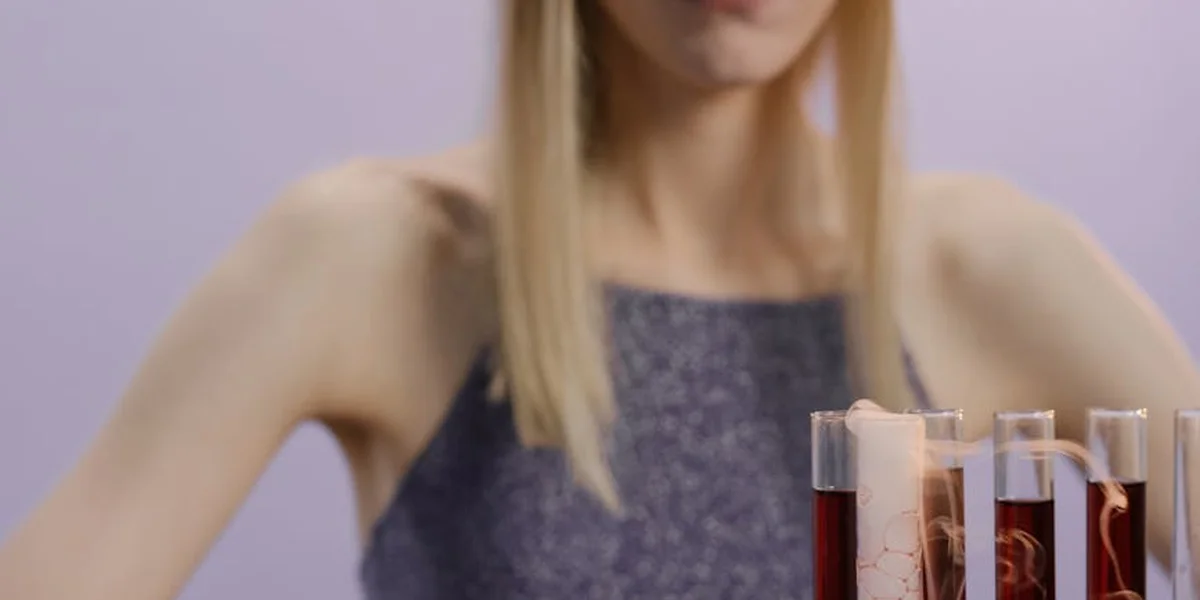 A softly focused image of a woman with arms raised against a light purple background, with several test tubes containing red liquid in the foreground.