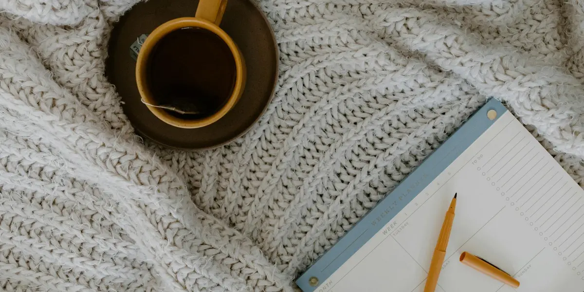 A cozy scene featuring a cup of coffee on a saucer, a pencil, and an open notebook resting on a chunky knit blanket, suggesting reflection and planning.