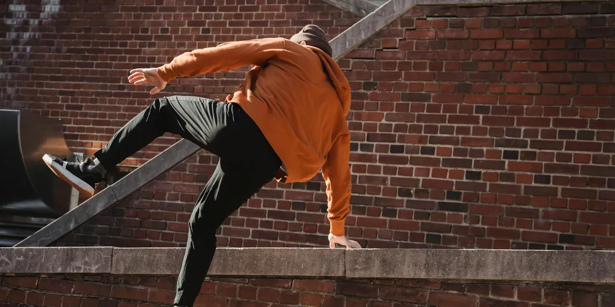 A person wearing an orange hoodie and dark pants leaps over a concrete ledge, with a brick wall in the background.