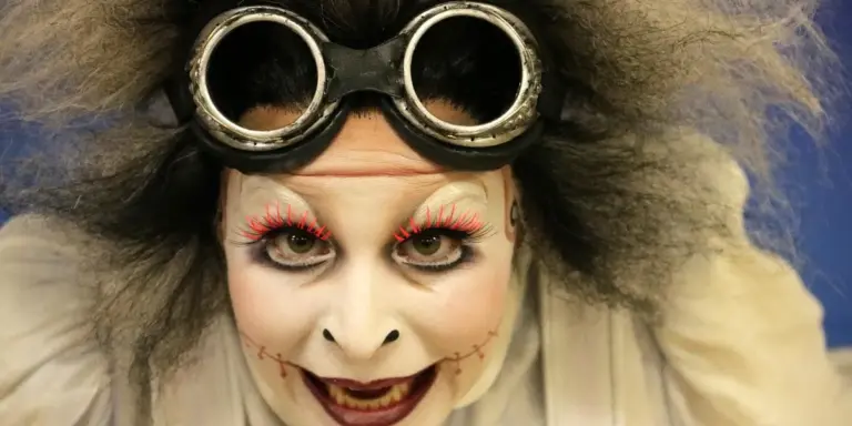 A person with wild hair and white clown makeup wearing round black goggles, creating a haunting, uncanny appearance.