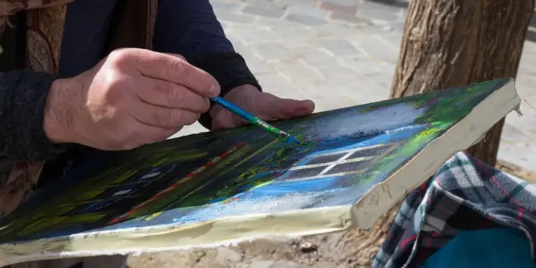 Close-up of a person painting on a colorful canvas outdoors, with a brush in hand and blue-green tones dominating the scene.