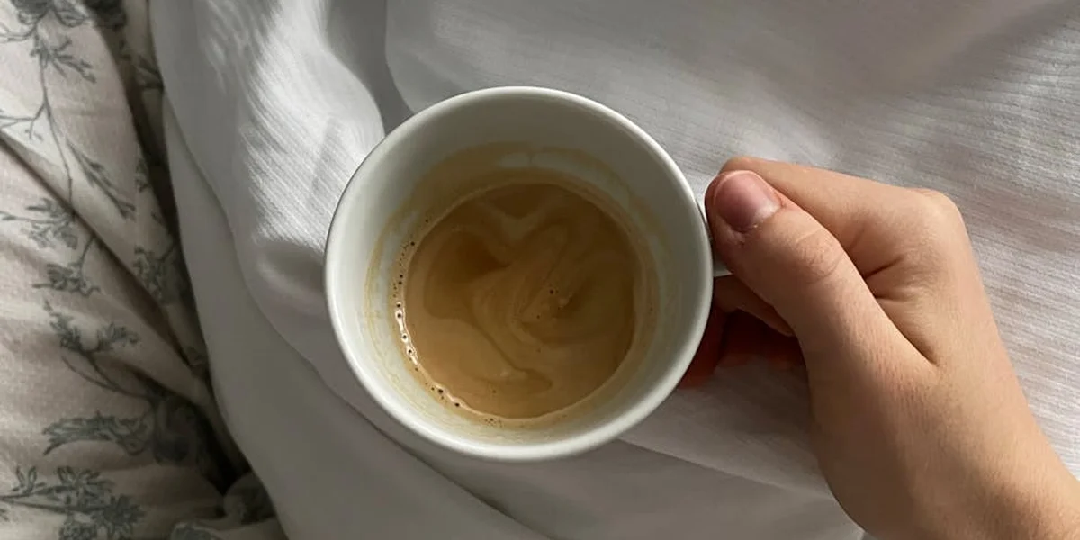 A hand holding a cup of coffee in bed with white sheets, suggesting late-night sleeplessness and a need for comfort.