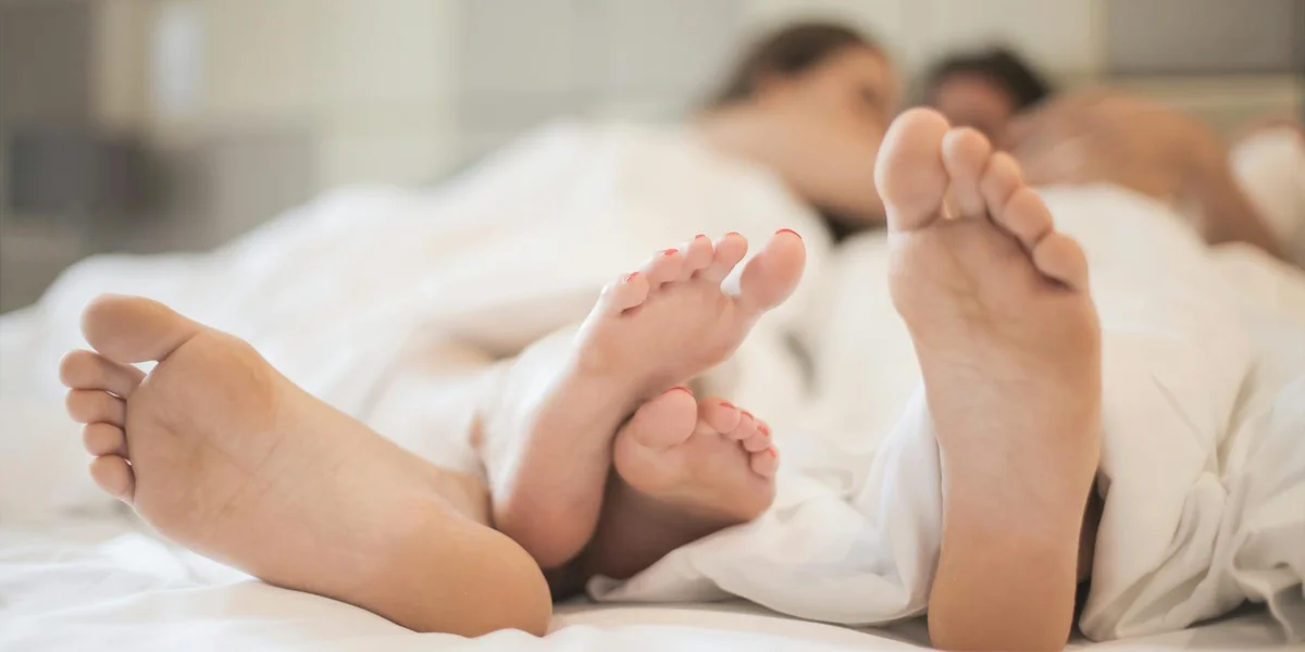 Close-up of bare feet in bed with a couple embracing under white sheets, softly blurred in the background.