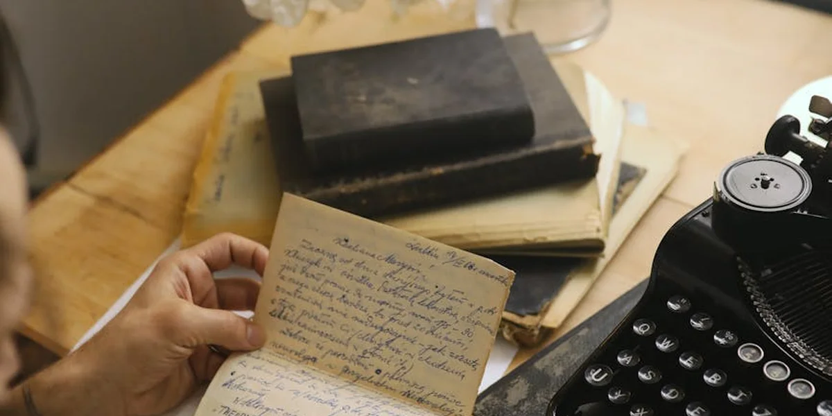 A hand holding a handwritten note over a cluttered desk with old notebooks and a vintage typewriter.