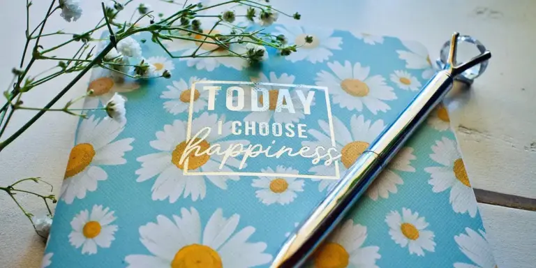 Blue notebook adorned with white daisies, a silver pen resting on it, and the phrase 'Today I choose happiness' printed on the cover.