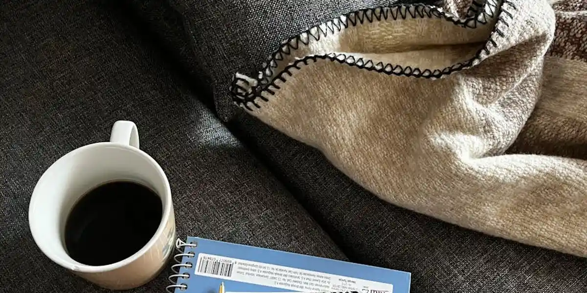 White coffee mug beside a small spiral notebook on a dark surface, with a beige blanket nearby.