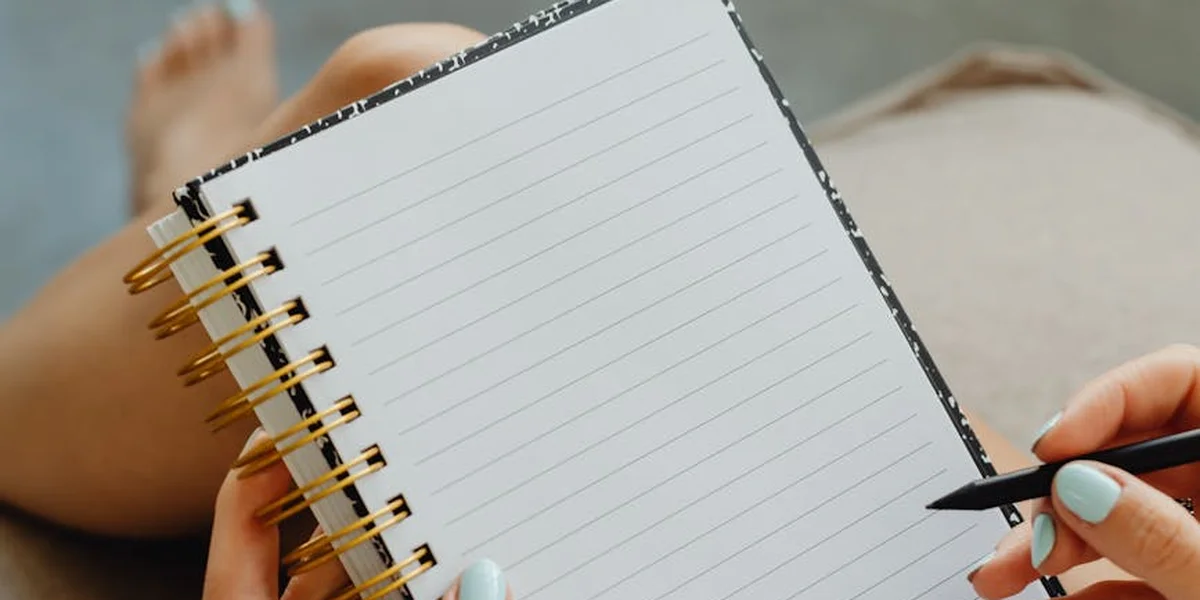 Close-up of an open lined notebook with a spiral binding; a hand with light blue nails holds a pen, while blurred feet rest in the background.
