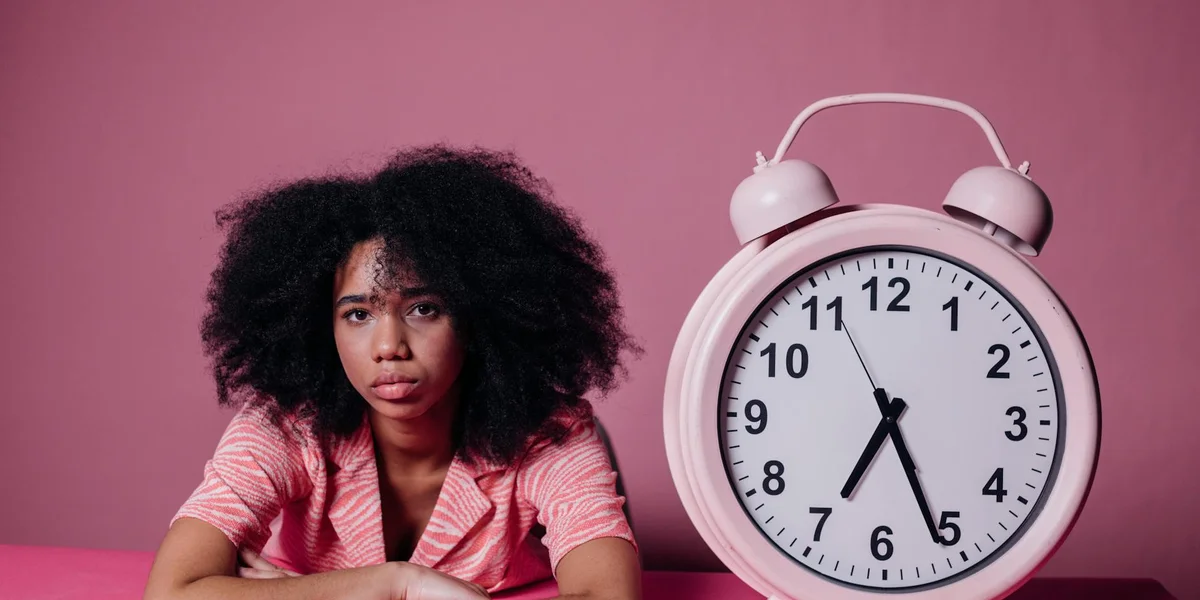 Person with curly hair seated beside a large alarm clock on a pink background, looking anxious about the time.