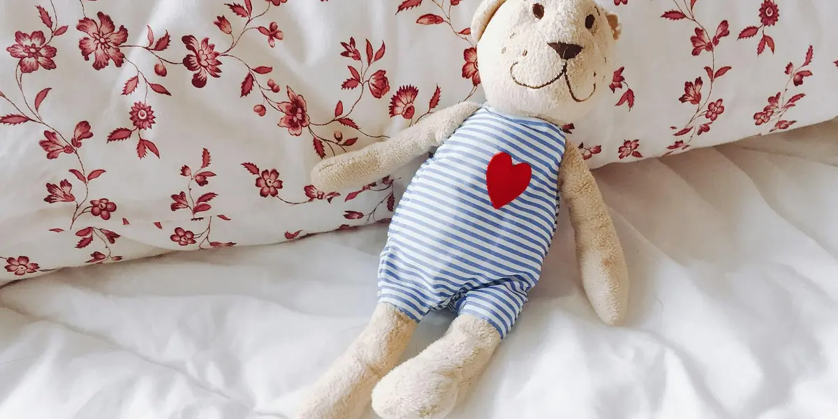 A plush teddy bear wearing a blue and white striped outfit with a red heart on its chest sits on a white bed with floral pillows.