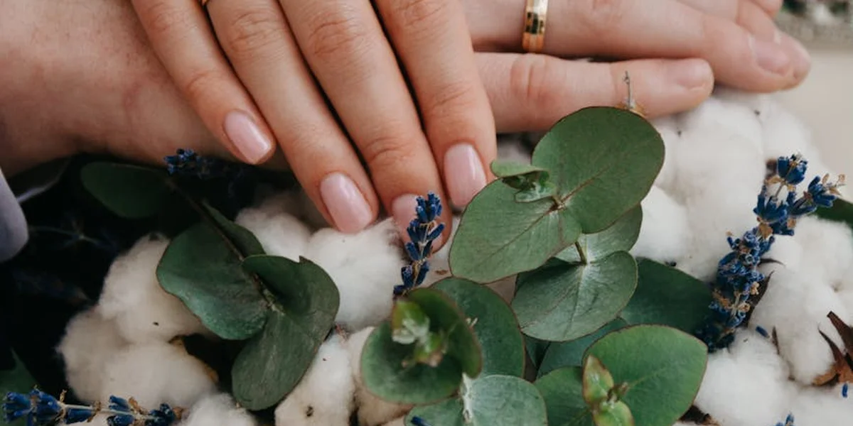 Close-up of a person's hands with a gold wedding ring resting on a soft white surface, surrounded by eucalyptus leaves and small purple flowers.