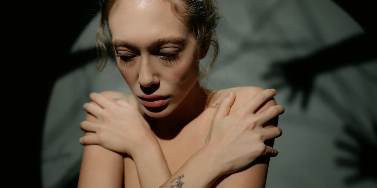 A woman stands with her arms wrapped around her shoulders, looking down with a pained expression.