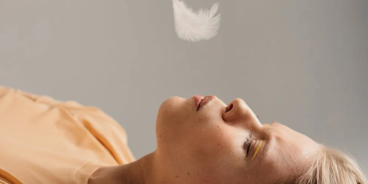 A close-up of a person lying on their back with eyes closed and head tilted, a soft white feather floating above, suggesting dream imagery and inner pain.
