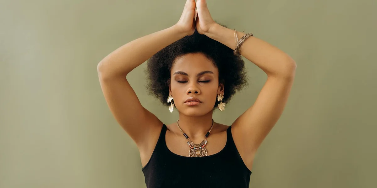 Person with eyes closed in prayer, hands pressed together above their head, wearing a necklace, symbolizing spiritual practice.
