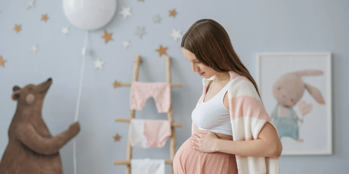Pregnant woman gently cradling her belly in a cozy, pastel-toned room with child-friendly decor.