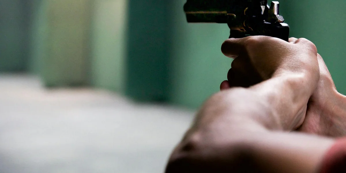 Close-up of a person’s arm gripping and aiming a handgun, with a blurred green background.