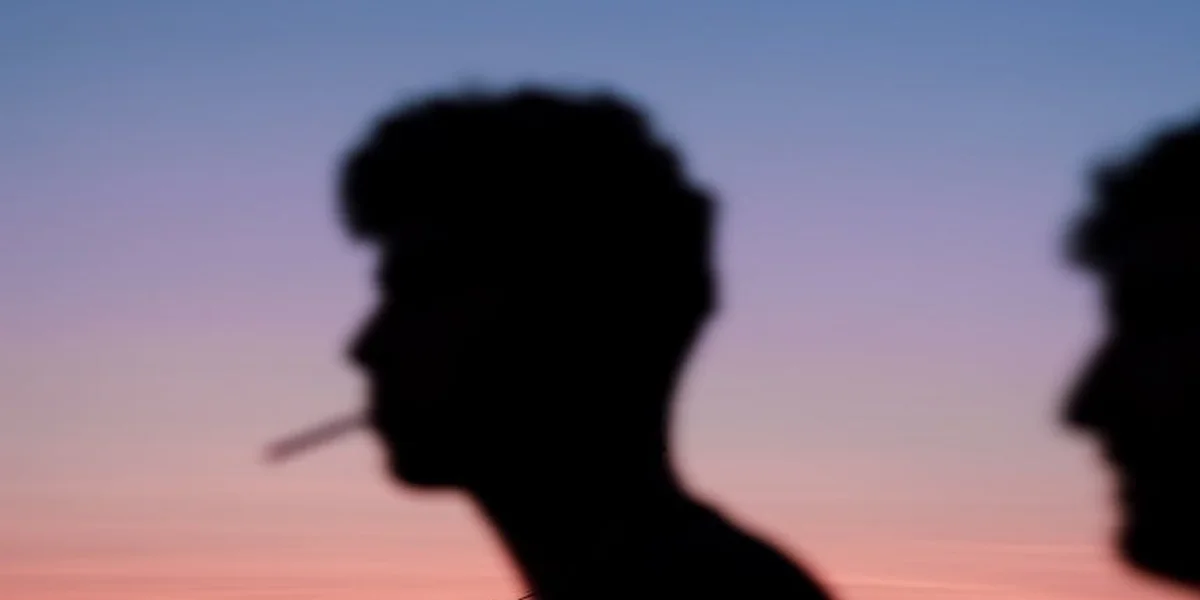 Silhouettes of two people at sunset, one smoking a cigarette, against a gradient sky.