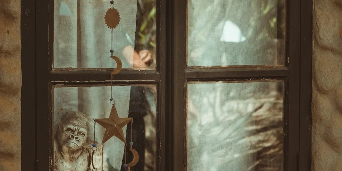 A window with a hanging arrangement of dream-themed symbols: a star, a crescent, and a small figurine, with soft light and greenery outside.