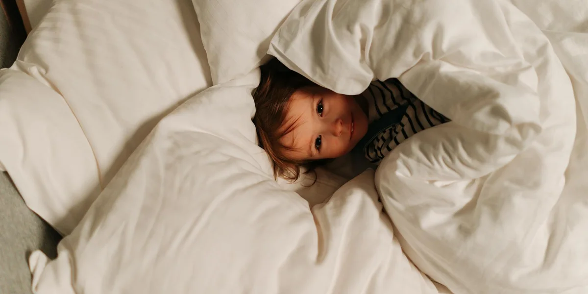 A young child peeks out from under a white comforter in a cozy bed.