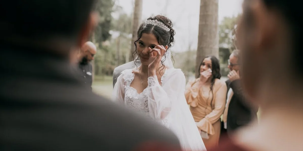 A bride in a wedding dress wipes away a tear during a ceremony, with guests visible in the background.