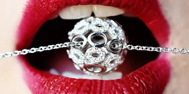 Close-up of red lips biting a silver, jewel-encrusted ball attached to a chain.