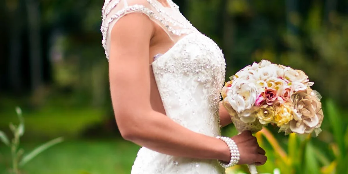 Bride in a white lace wedding dress holding a bouquet outdoors; close-up on lace, beading, and pearl bracelet.