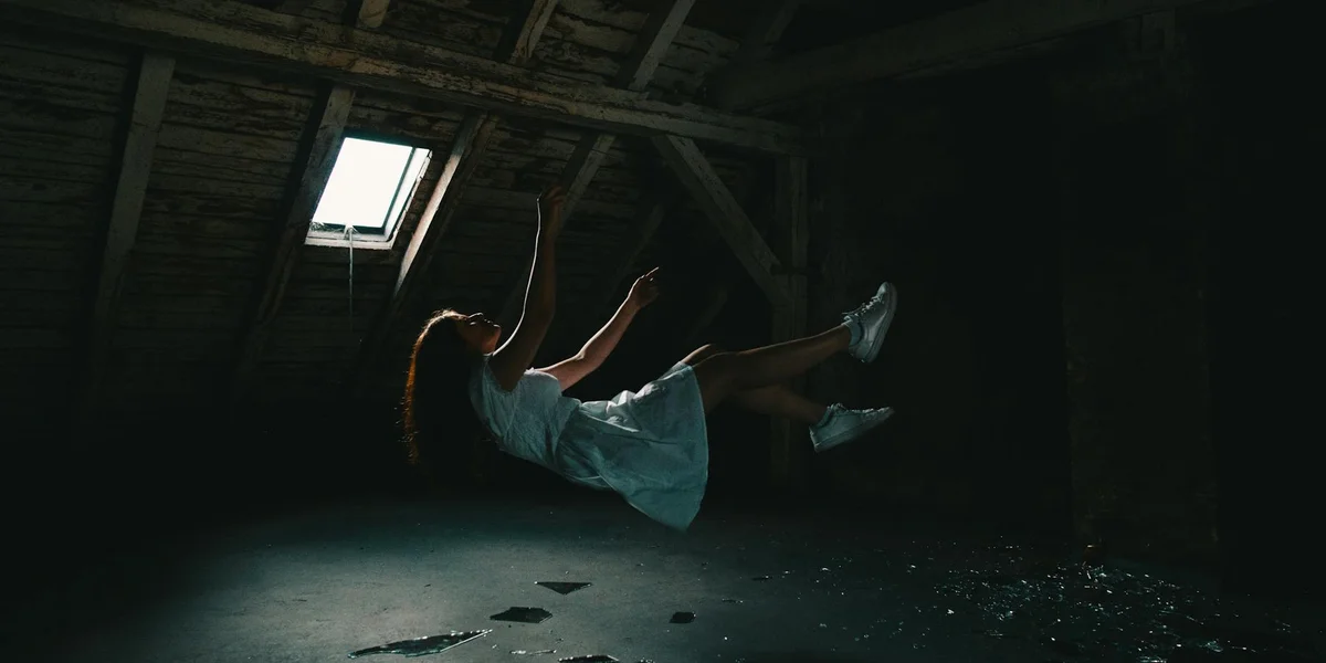 A person in a light dress floats mid-air in a dark attic, illuminated by a shaft of light from a small skylight.