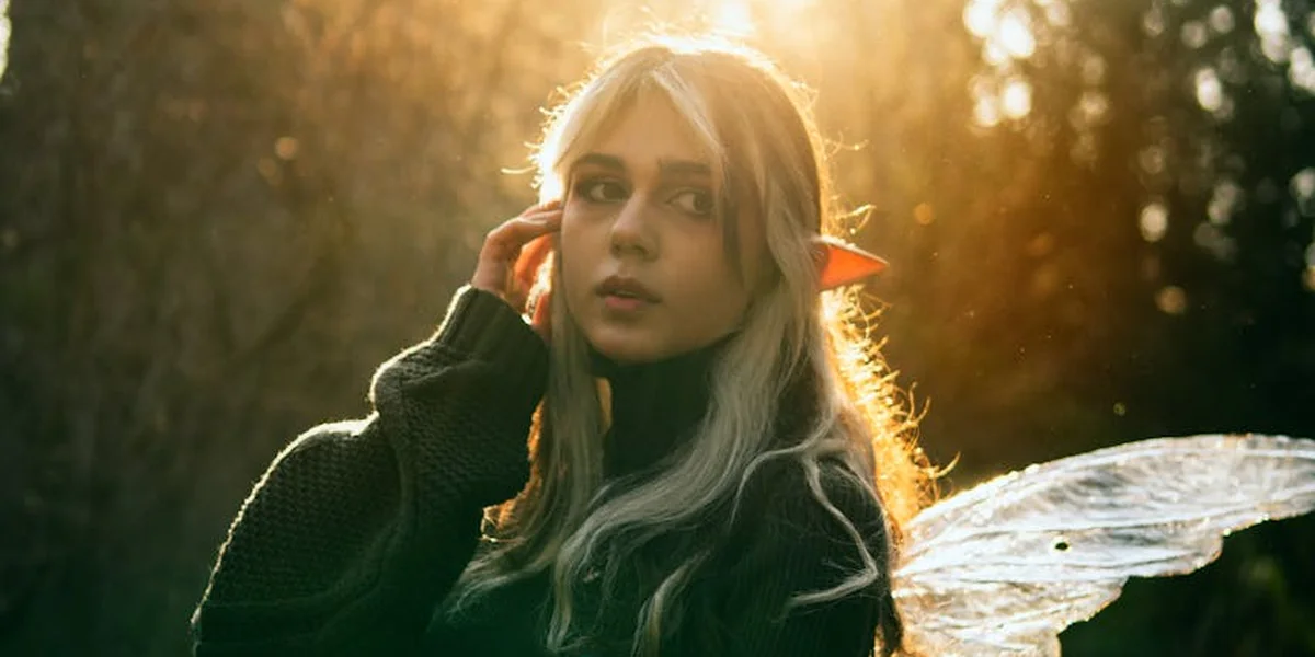 A young woman with long blonde hair stands outdoors during golden hour, wearing a dark sweater and looking slightly to the side.