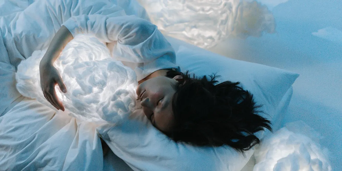 Person sleeping in a bed with white bedding under blue-toned lighting, evoking dreaming and introspection.