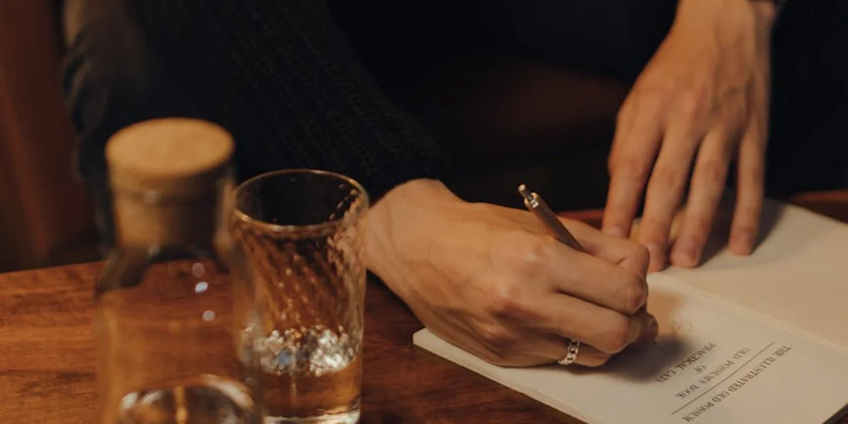 Close-up of a person writing on a sheet of paper at a wooden table, with a glass of water nearby.