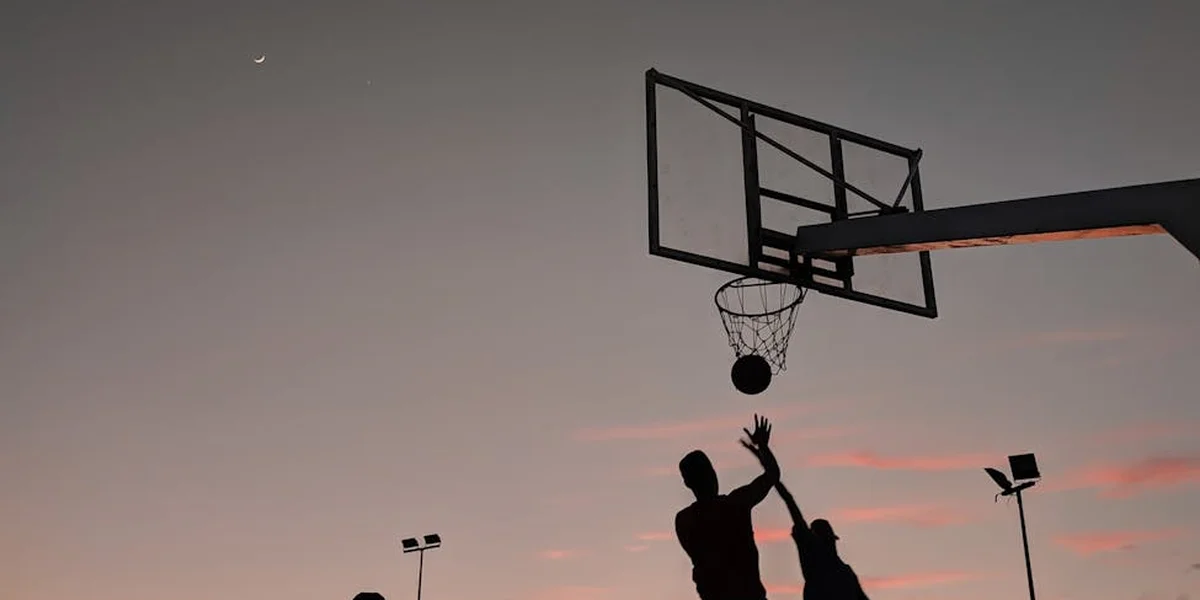 Two players silhouette against a sunset, reaching toward a basketball hoop, symbolizing effort and resilience.