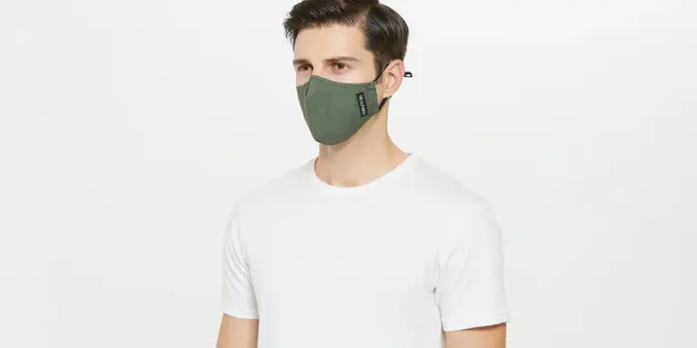 A person wearing a white t-shirt and a green face mask stands against a plain light background.