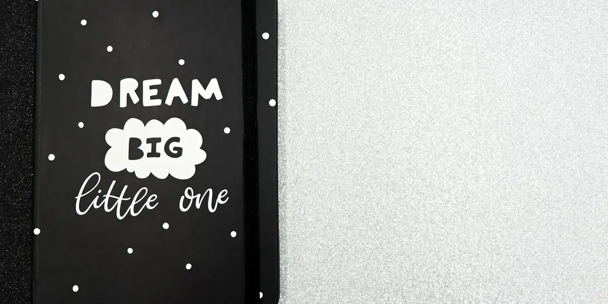 Close-up of a black notebook cover showing the words 'DREAM BIG' in bold white letters and 'little one' in cursive, surrounded by small white dots.