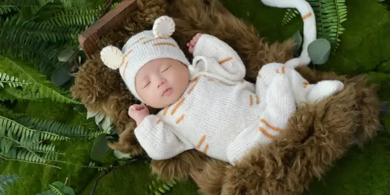 Newborn baby sleeping in a white knitted outfit inside a brown furry-lined basket, surrounded by green ferns.