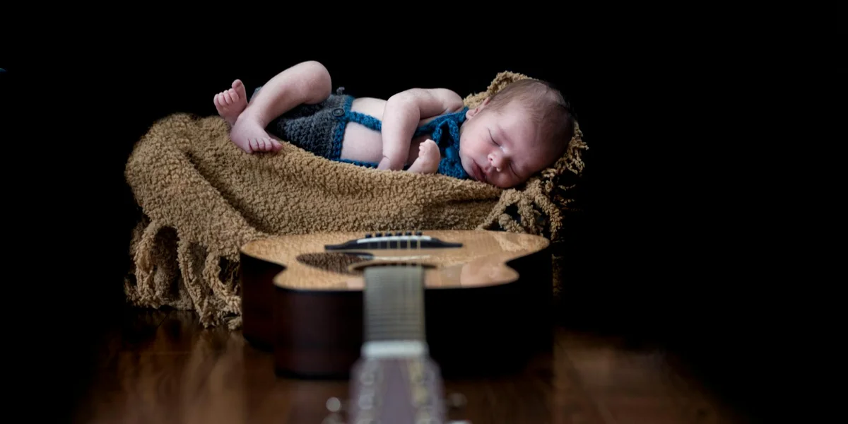 Newborn baby sleeping on a textured brown blanket with a guitar in the foreground.