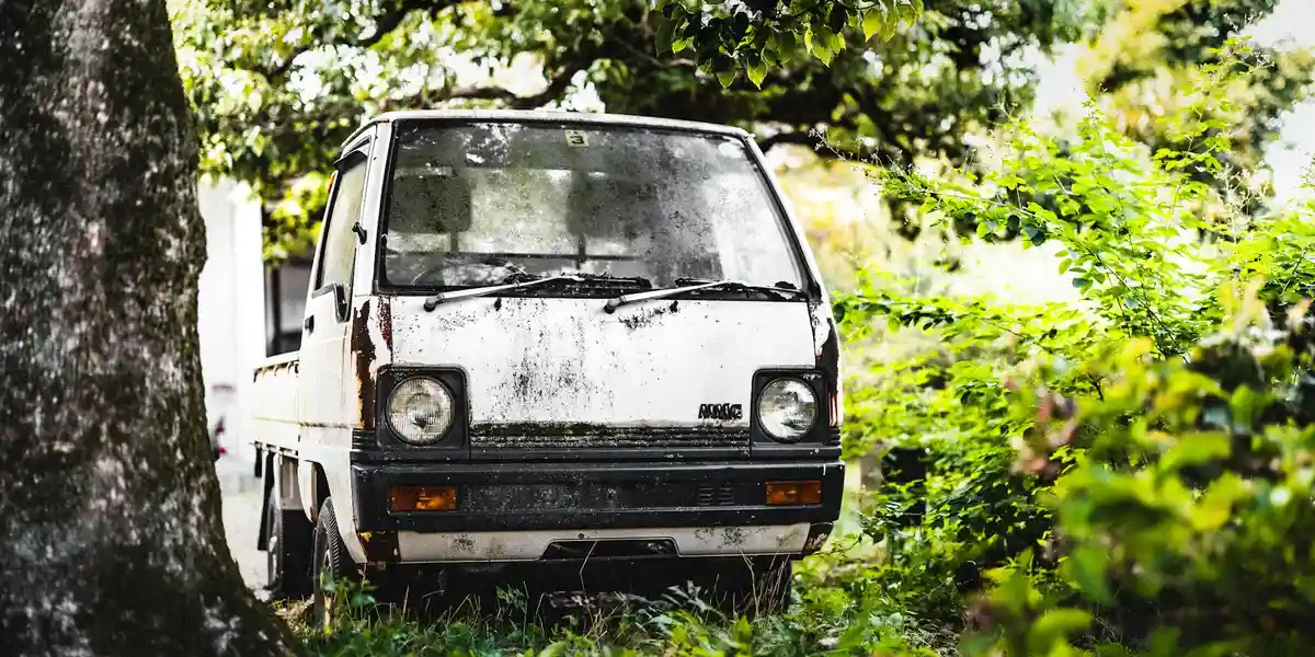 A small white van parked among dense green trees, suggesting stillness and a pause within a dream-city landscape.