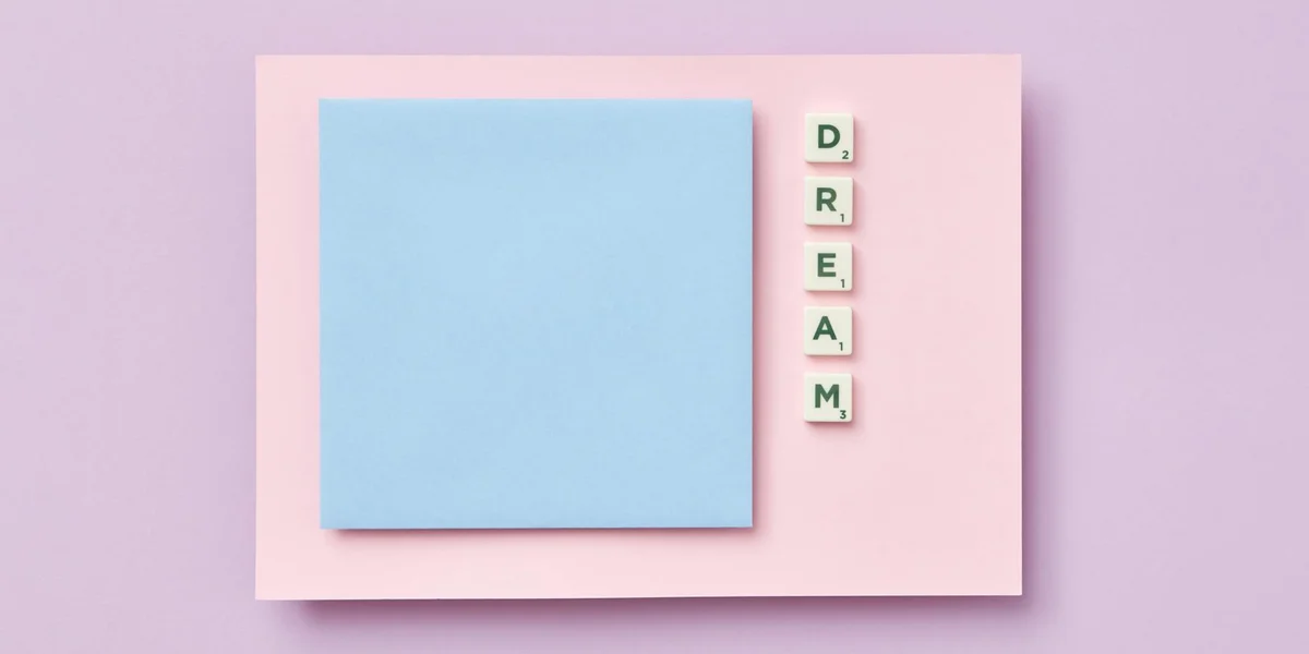 Pink card on a lavender background with a blue sticky note and Scrabble-style tiles spelling DREAM arranged vertically to the right.
