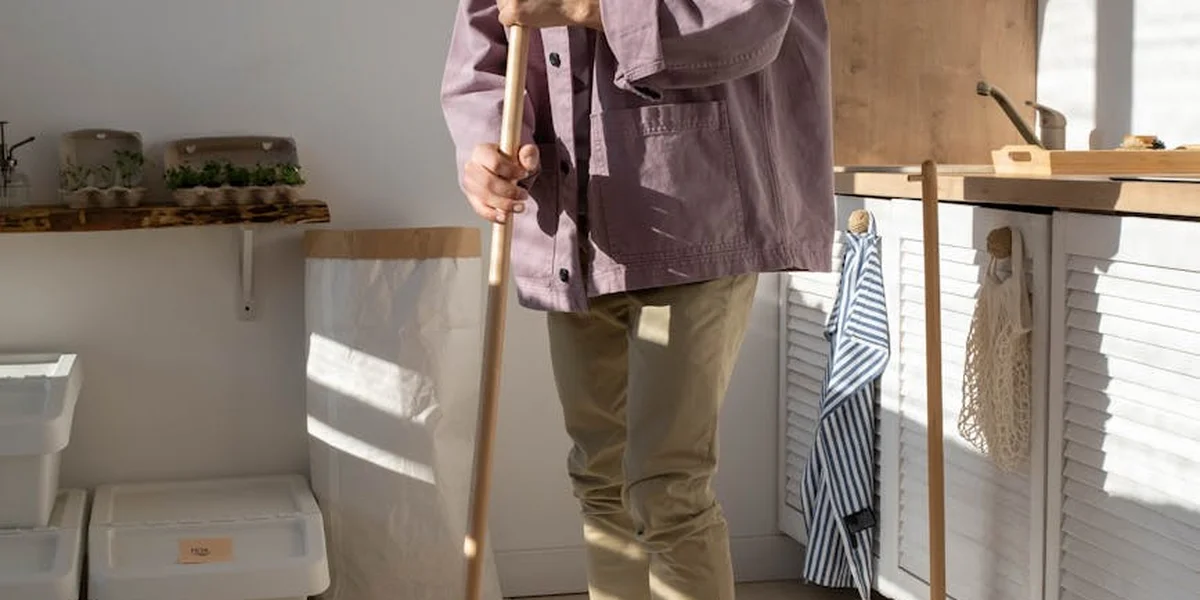 A person cleaning with a broom in a bright, sunlit kitchen; shelves, towels, and cleaning supplies are visible in the background.