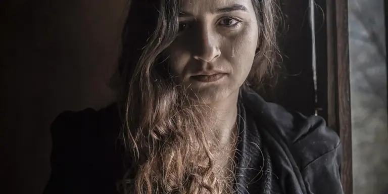 A somber woman sits near a window in a dimly lit room, gazing downward with a serious, contemplative expression.