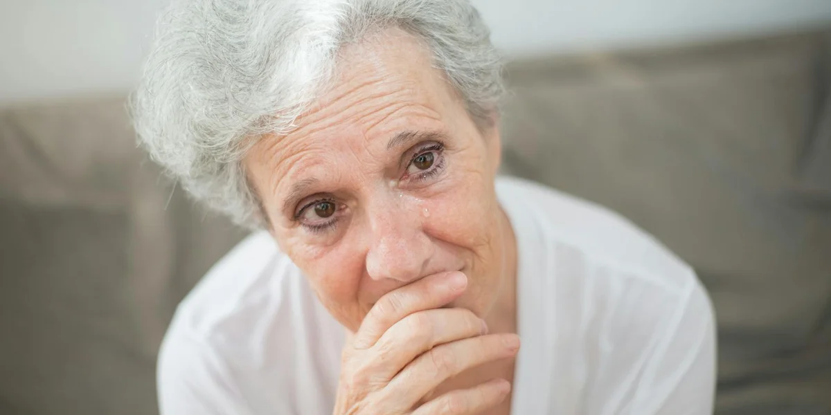An older woman with short gray hair sits on a sofa, her hands gently covering her mouth as she looks pensive and distressed.