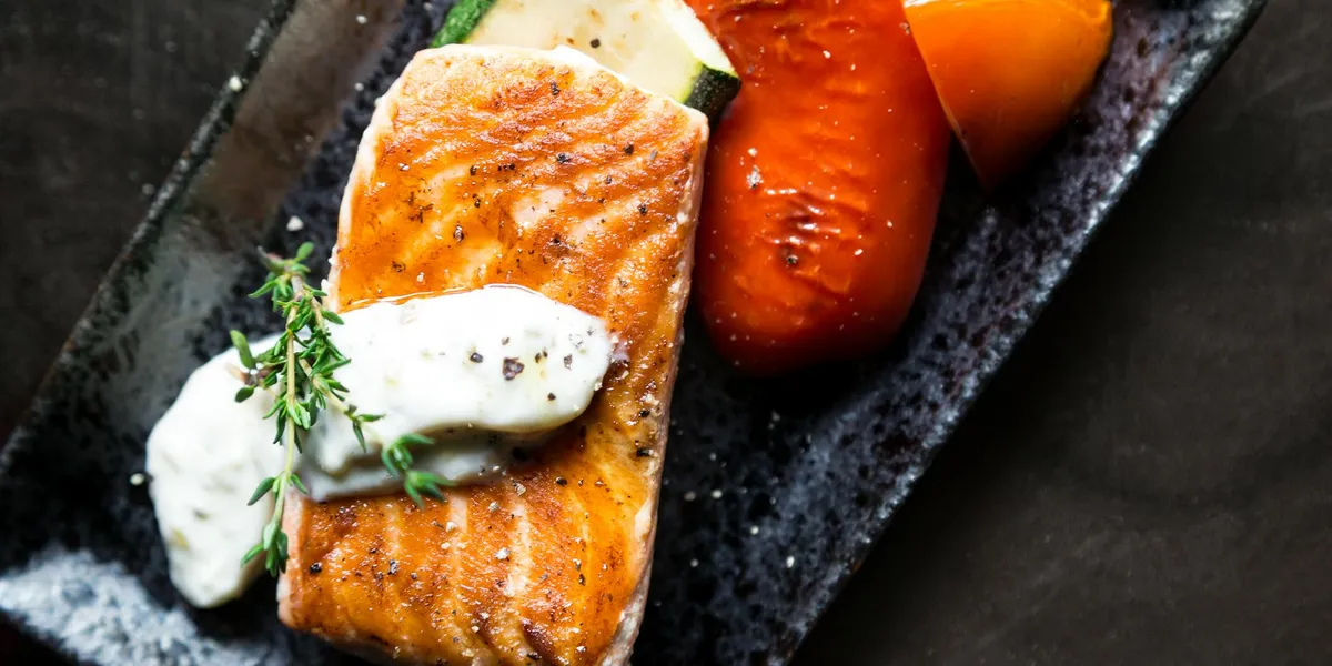 Close-up of a plated salmon fillet with herb garnish and roasted vegetables on a dark plate, conveying nourishment and mindful eating.