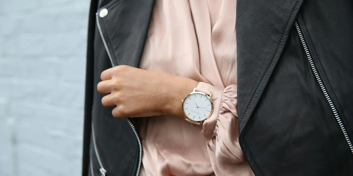 Close-up of a person wearing a pale pink blouse under a black leather jacket, with a white-faced wristwatch featuring a gold case.