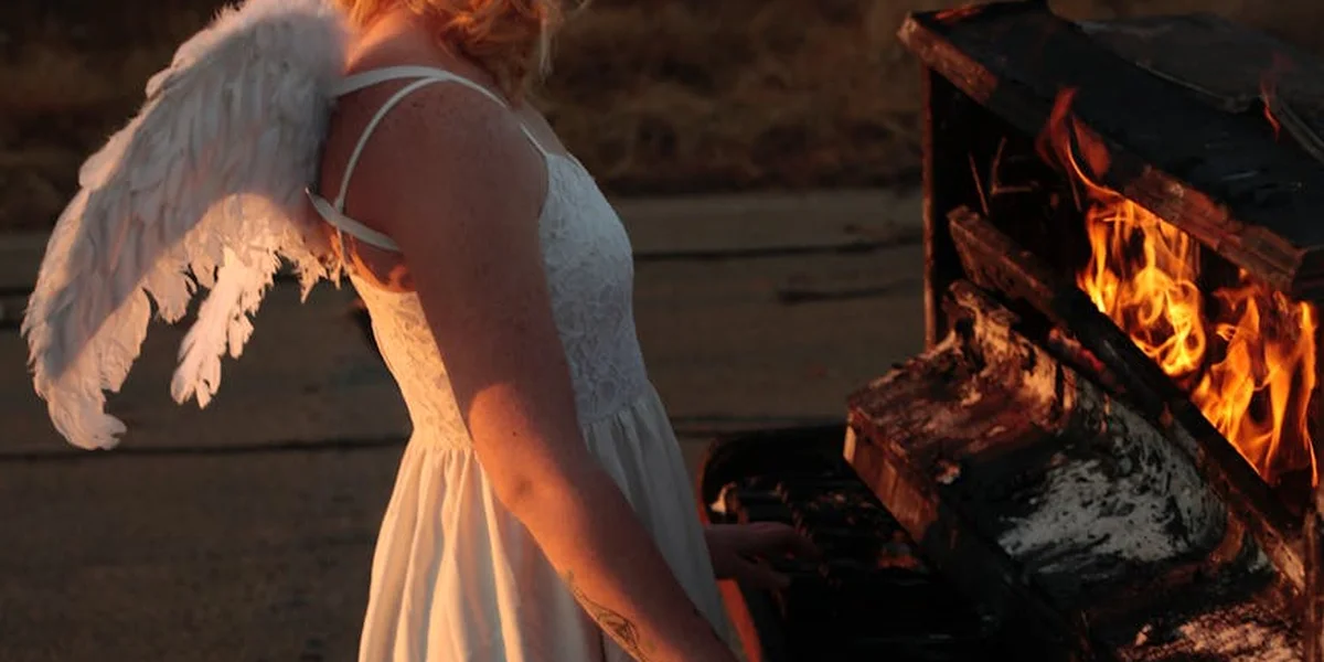 A figure wearing a white lace dress and angel wings stands beside a blazing fire, symbolizing flight and transformation.