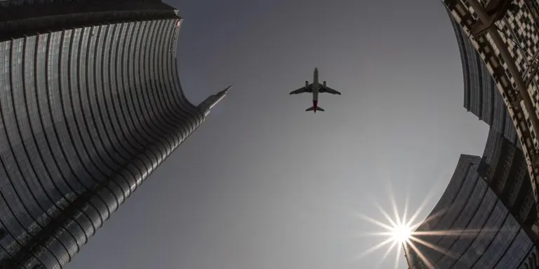 A plane flies overhead between tall city skyscrapers, with the sun creating a bright starburst in the clear sky.